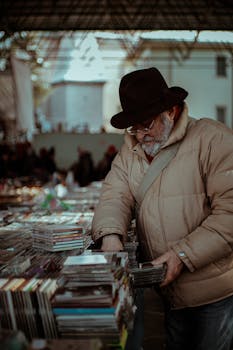 An elderly man wearing a hat and glasses browses CDs at an outdoor market in Ankara, Türkiye.