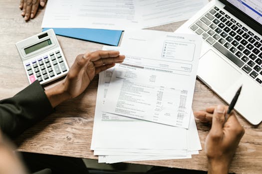 Hands holding financial documents with calculator and laptop on office desk, business analysis scene.