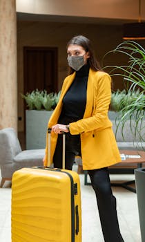 Woman in yellow coat and mask holding suitcase, embracing new normal travel.