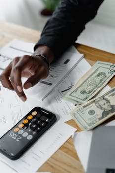 A person calculating expenses using a smartphone and counting cash bills on a wooden table.