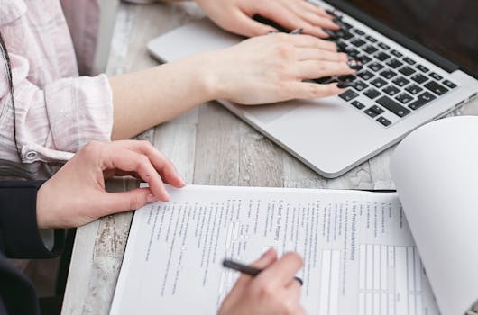 Close-up of hands typing on a laptop and reviewing business documents, focused on finance and legal tasks.